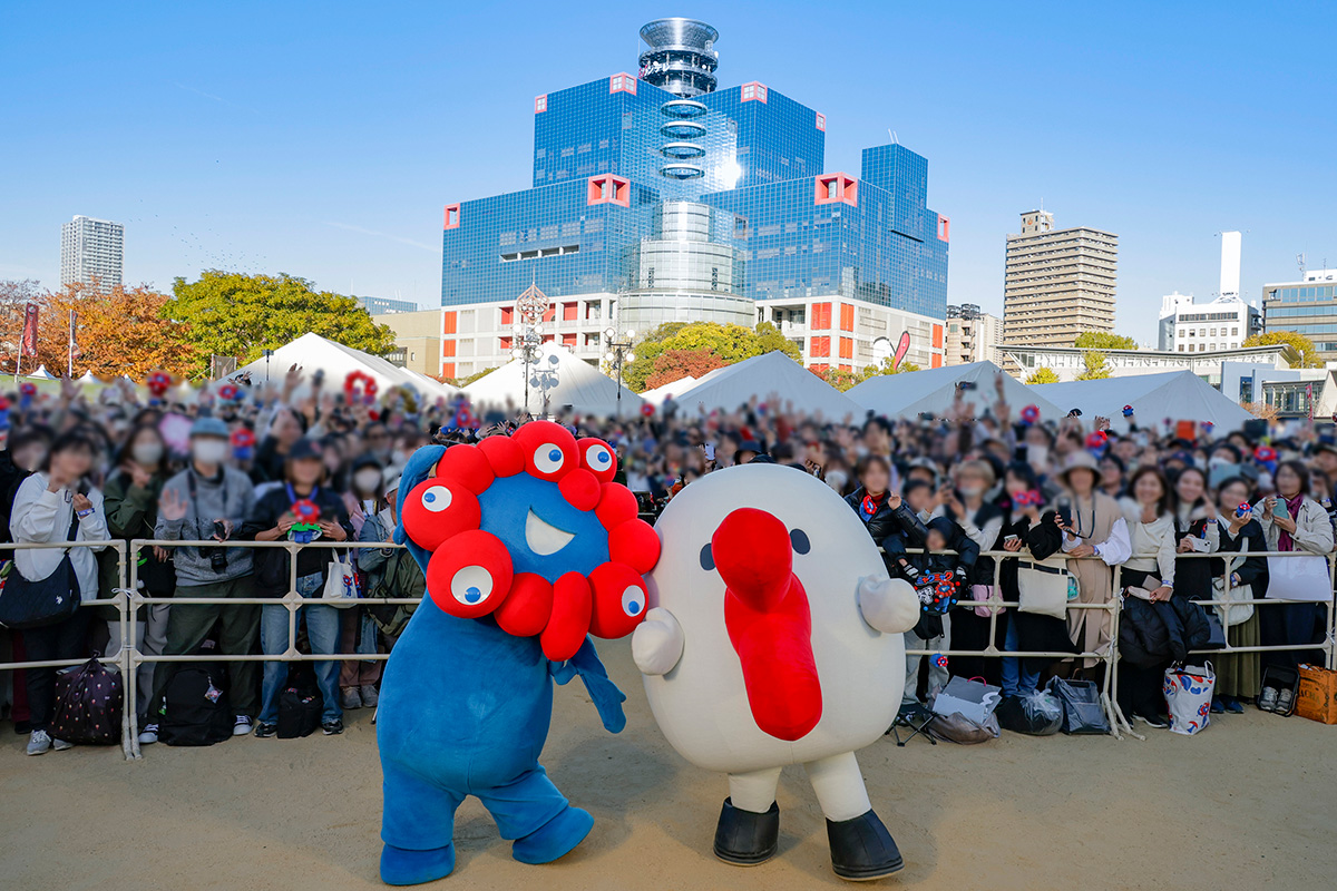 観客と記念撮影をするミャクミャクとハチエモン、奥にはミャクミャク色のカンテレ社屋（2025年11月23日・大阪市北区）