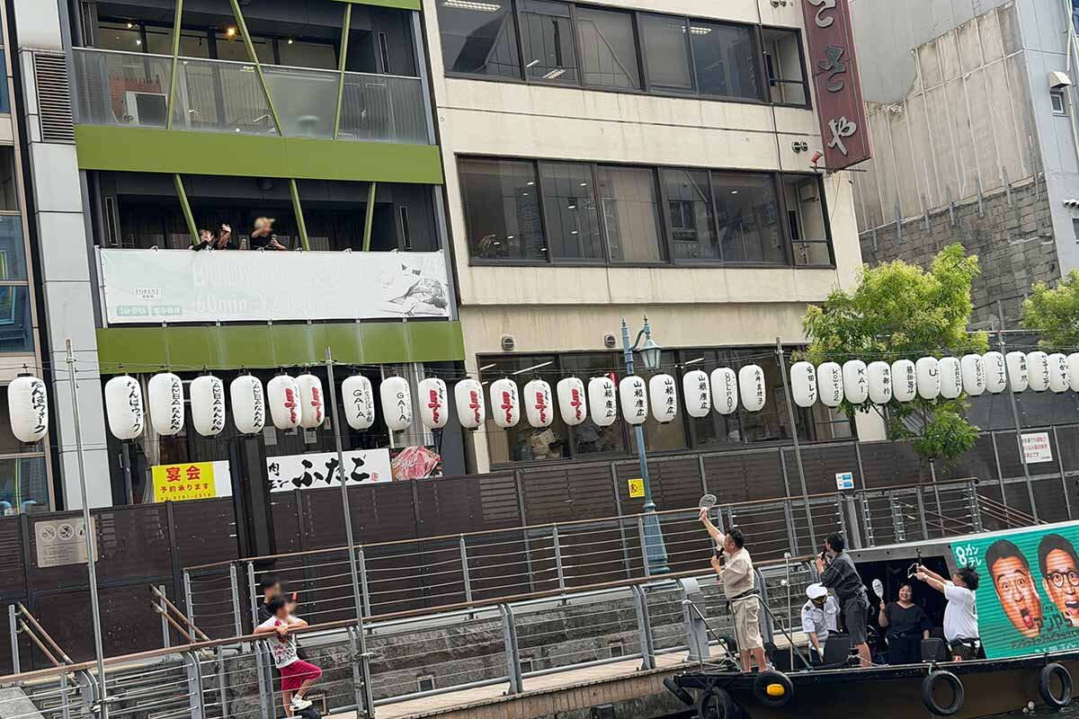 ビルから手を降る地元の人たちに船上から応えるアンタッチャブルの2人（2026年8月・大阪市中央区）