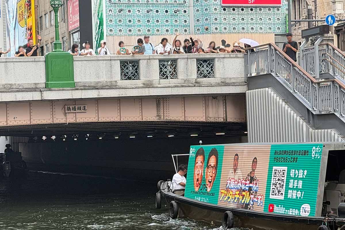 沿岸や橋の上からも声援を受けるアンタッチャブルの2人が乗るクルーズ船（2026年8月・大阪市中央区）